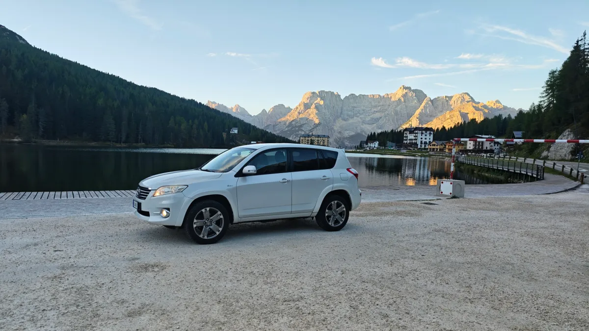 Toyota RAV4 branco estacionado em frente ao Lago Misurina nas Dolomitas ao entardecer com picos nevados ao fundo.